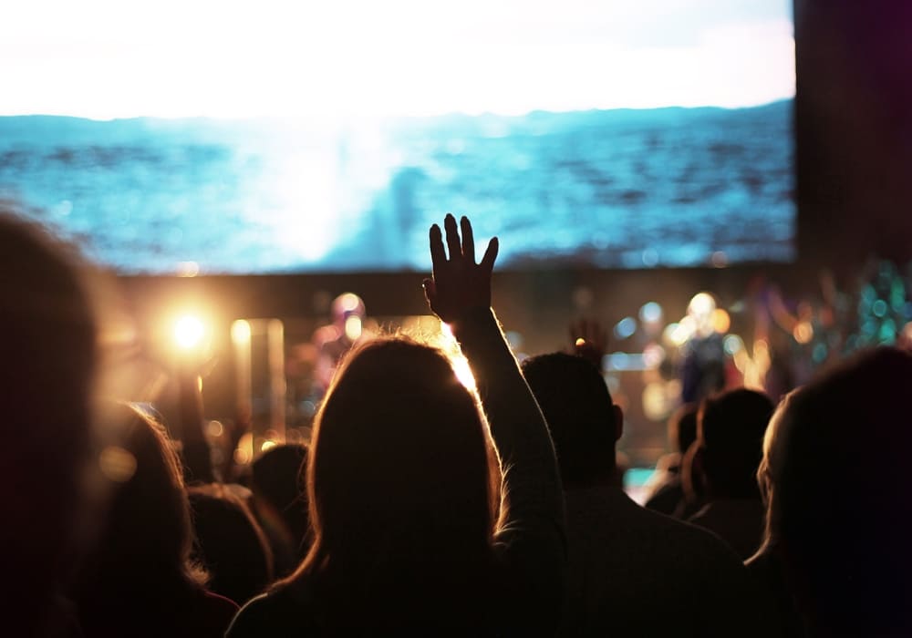 A congregation worshiping with hands raised in praise, illuminated by soft lights, with a serene ocean scene displayed on a large screen in the background.