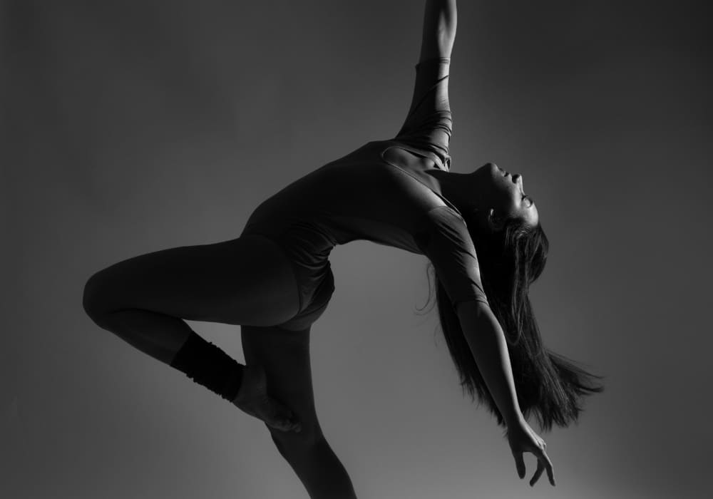 A black-and-white photograph of a female dancer striking a graceful pose, arching her back with one leg bent and an arm extended upward, embodying elegance, strength, and passion.