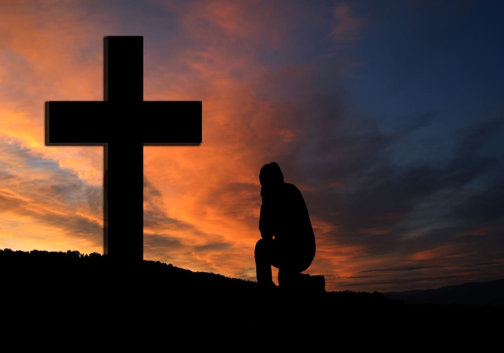 A silhouetted figure kneeling in prayer beside a cross at sunset, set against a dramatic and colourful sky.