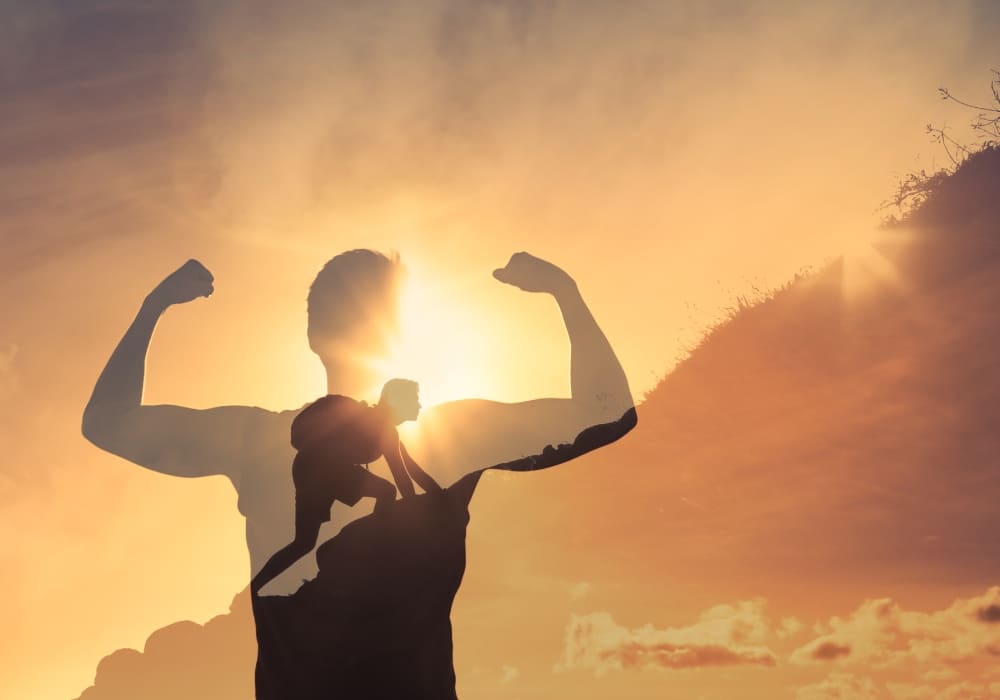 A double-exposure image blending a person flexing their arms in strength with a silhouette of a climber scaling a steep hill, set against a radiant sunrise or sunset sky.