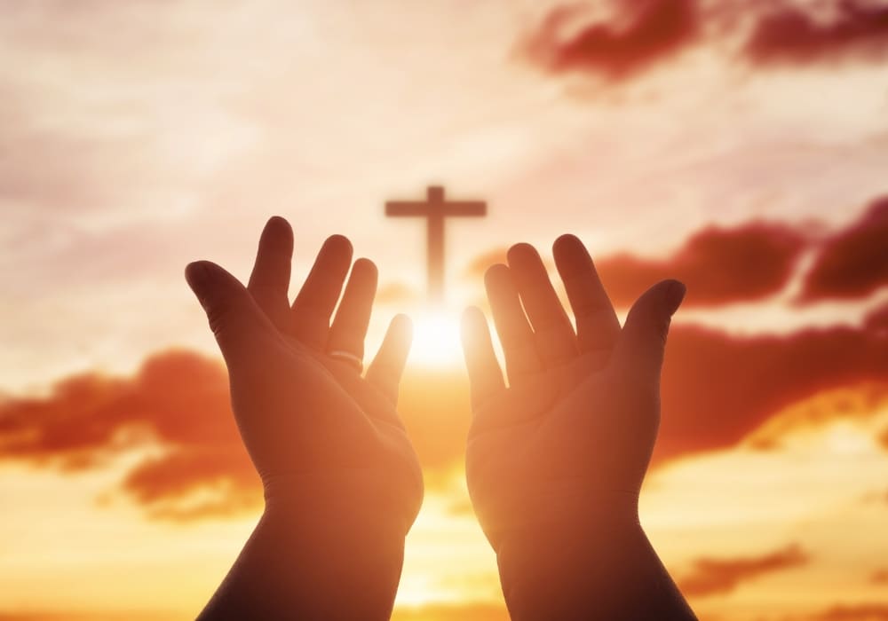 Hands raised in prayer towards a glowing cross in a vibrant sunset sky, symbolizing faith and trust in God's provision.