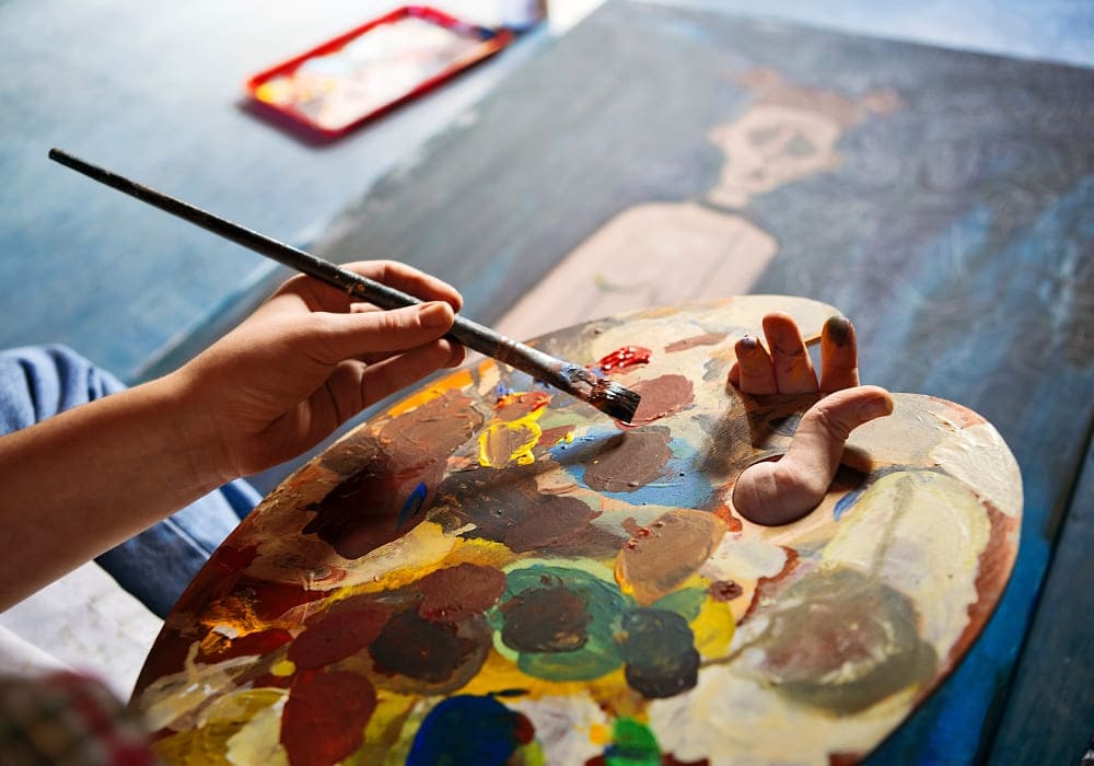 An artist's hand holds a paintbrush and palette, adding colours to a canvas, symbolising creativity, patience, and the journey of self-expression.