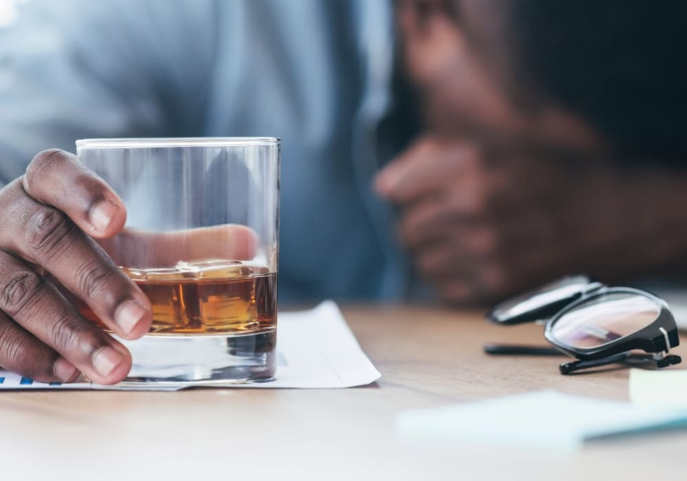 clemhholloway.com-Stressed man holding a glass of whiskey at his desk, with eyeglasses beside him, symbolizing workplace burnout.