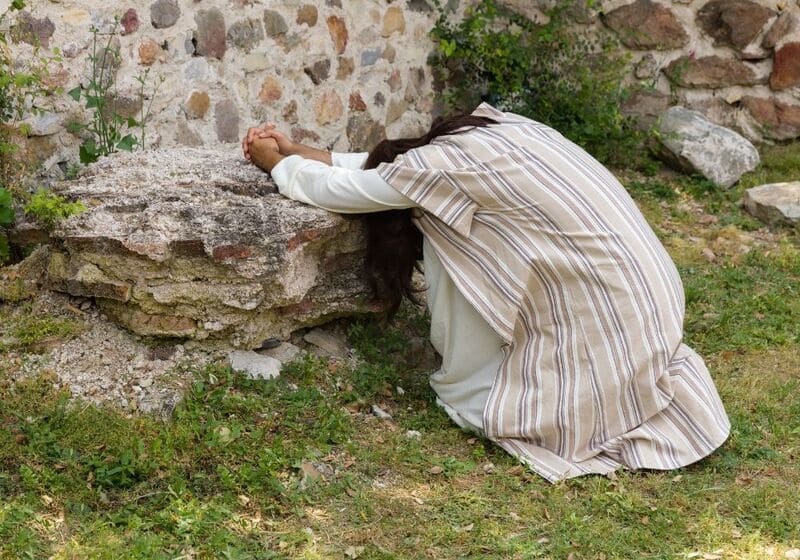 Alt Text: A person in biblical-style clothing kneeling in prayer against a stone wall, symbolising deep contemplation and spiritual struggle.