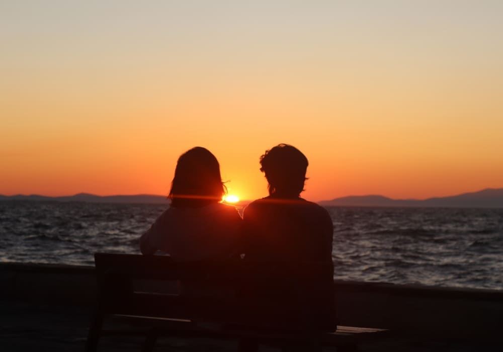 Two people sitting on a bench watching a sunset by the sea.